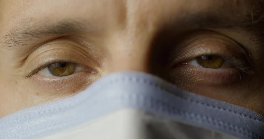A brown eyed man wearing a mask over his nose and mouth looks straight ahead and blinks