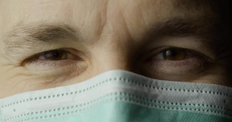 A man wearing a mask covering his nose and mouth looks straight ahead and his face lifts as he smiles under the mask