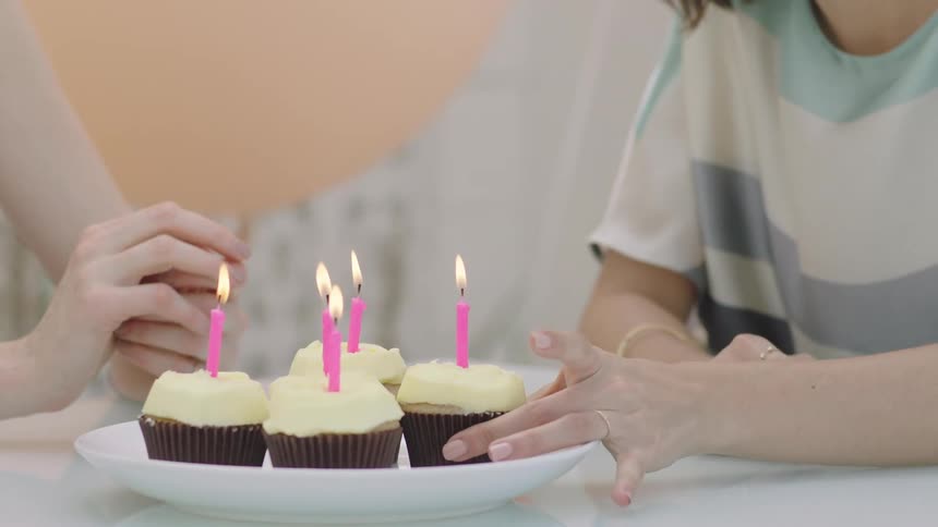 Two white women sit at a table and one of them lifts a cupcake with a candle on it