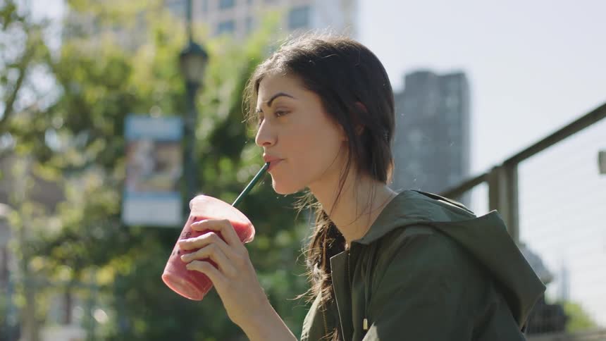 A woman sitting outdoors in a sitting drinking a red colored drink from a plastic cup