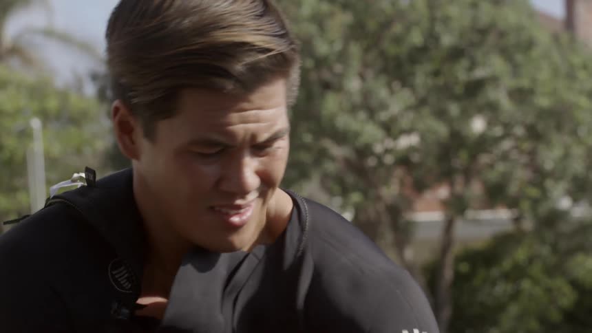 A young man putting on a wet suit as he squints in bright sunlight