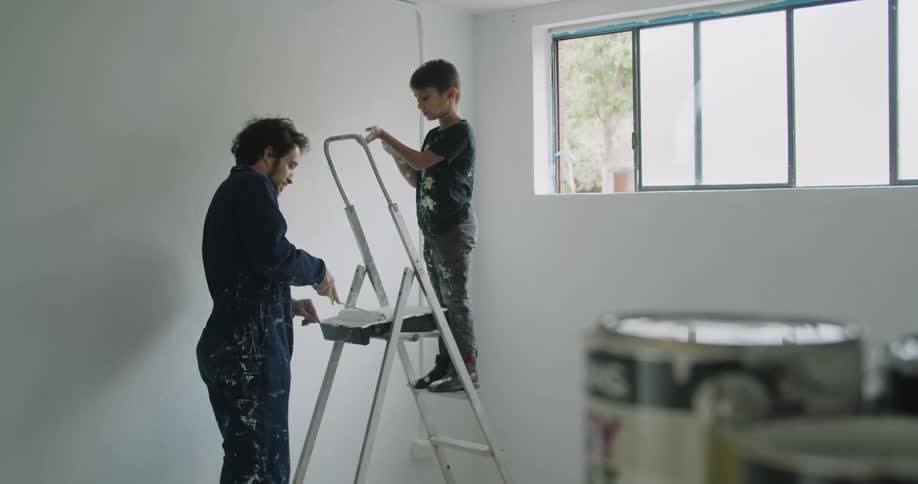 A boy stands on a step on a ladder while his dad loads a roller with paint from a tray to paint a room