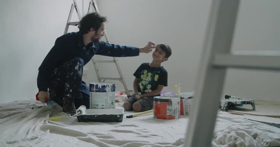 A dad puts paint on his sons face as they kneel on a tarp taking a break from painting