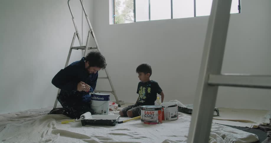 A white man and a child sit on the floor of a room that is about to be painted
