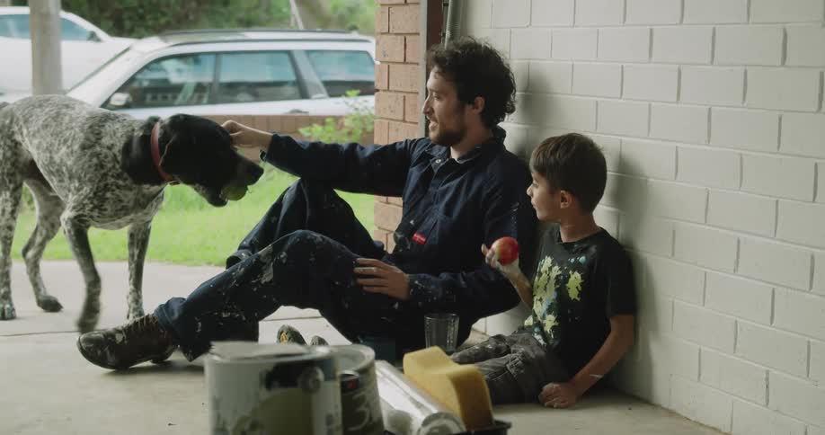 Man and boy sit on ground eating and playing with dog