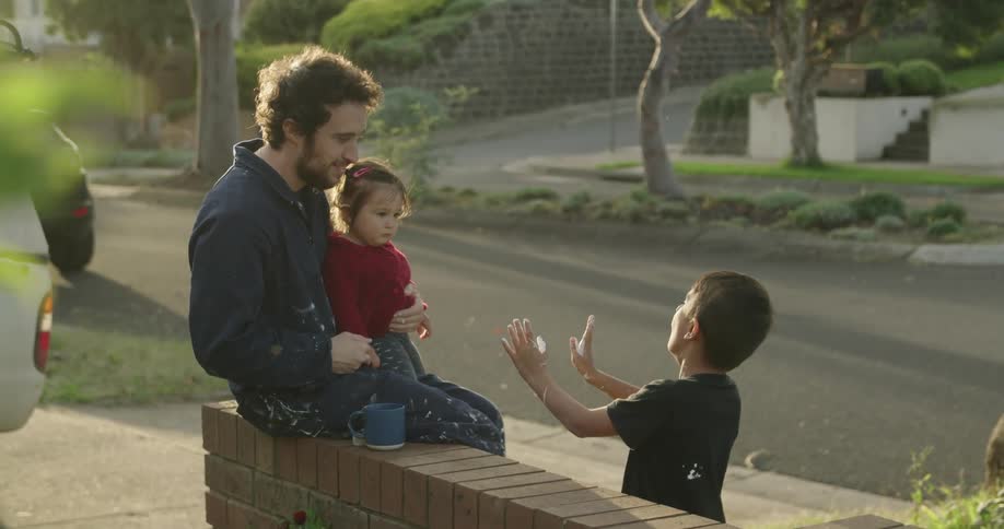 A young boy plays with bubbles and shows his father and sister as a vehicle passes by their house