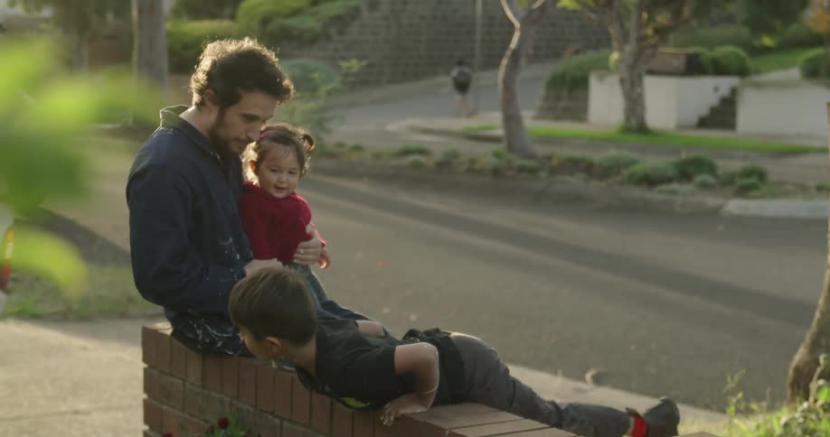 A man sitting on a low brick wall holding a little girl in his lap while a boy plays on the wall next to him