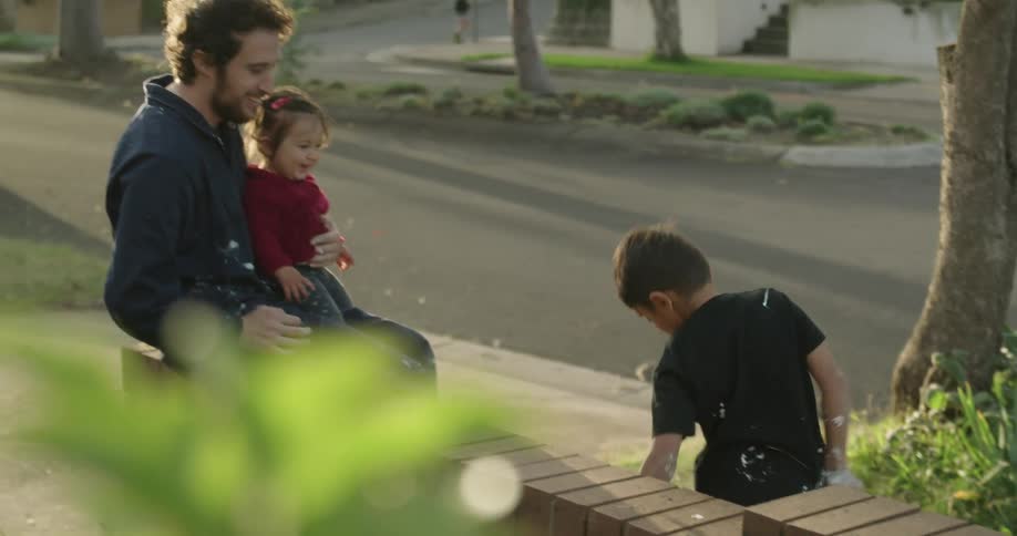 Father holding his daughter looks at his son playing on the pavement in the neighbourhood