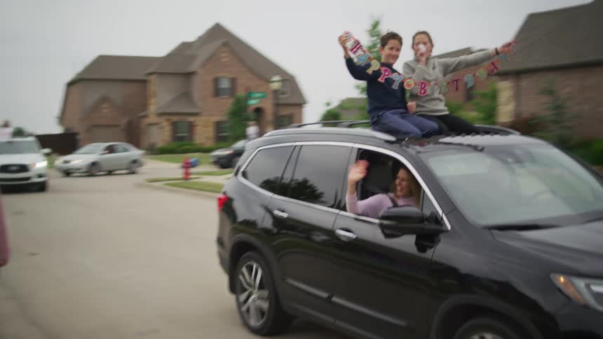 Children hold a happy birthday banner as they sit on the roof of a car passing through a neighborhood