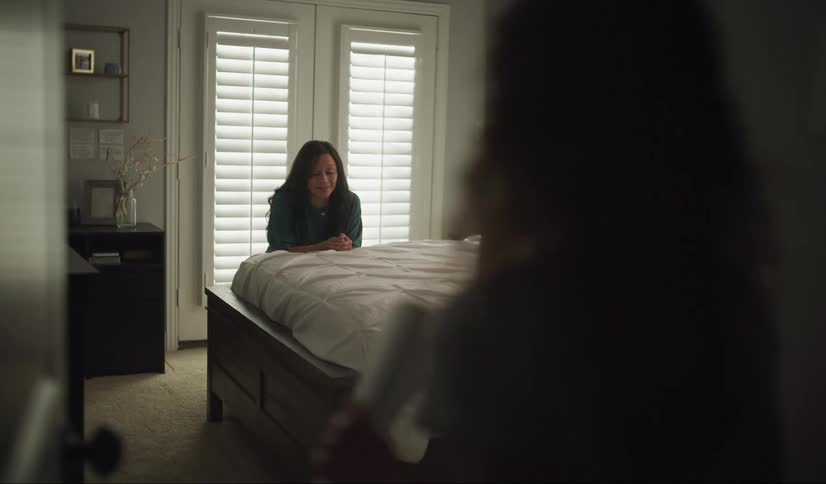 A woman kneels at her bedside in a prayer when a young girl walks to the bedroom doorway and watches.
