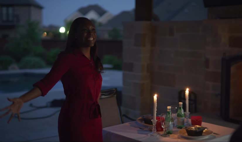 A black woman surprises her husband with a candlelight dinner of Chinese takeout on the back patio at dusk. They hug each other.