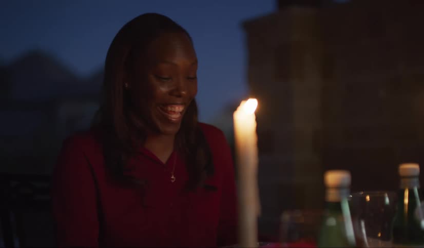 A black woman laughs while having drinks outdoors at night by candlelight