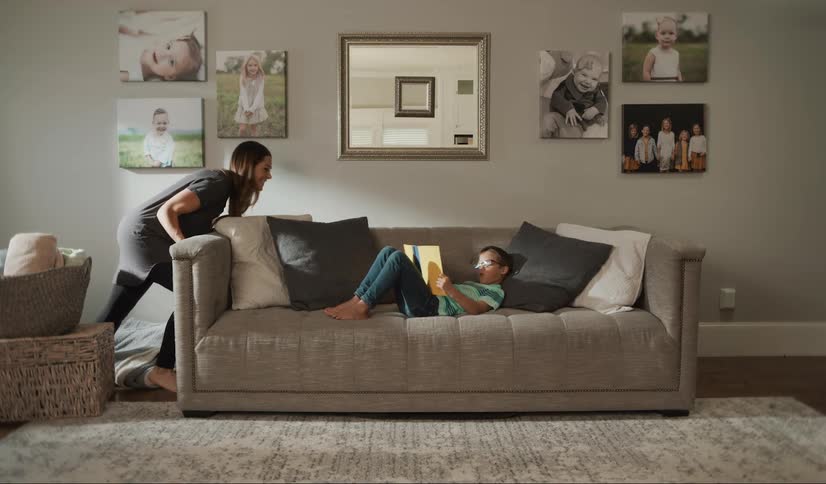 A white woman moves a couch across living room while a kid lays on it reading a book