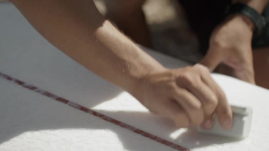 A man wearing sunglasses waxes a surfboard