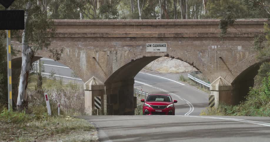 A couple traveling in a red car drive on a rural road under a bridge on a sunny day