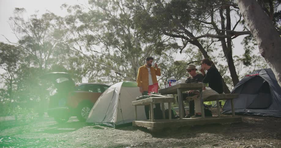 Two men sitting on a picnic table as another man stands as they drink tea with tents and a vehicle in the background