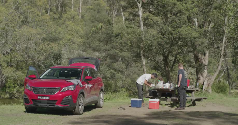 Two men pull food from cooler to place it on picnic table near parked car