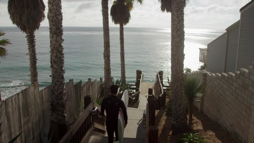 A man and woman, who are carrying surfboards, descend stairs that lead to the beach