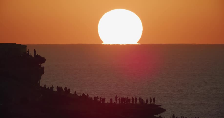 A sun sits on the horizon edge of the ocean with a crowd watching
