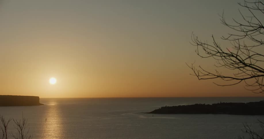 The sun sets over the ocean near a small island with a tree in the center