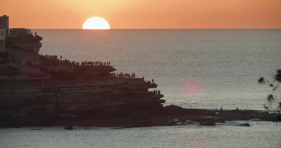 A large group of people line the shore and watch the sun set
