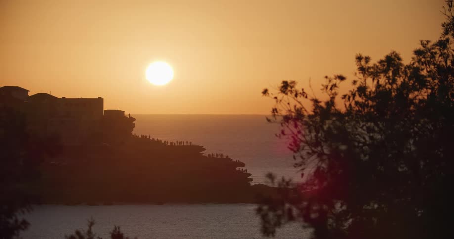 Sun hovers in orange sky casting building and promontory into silhouette against ocean