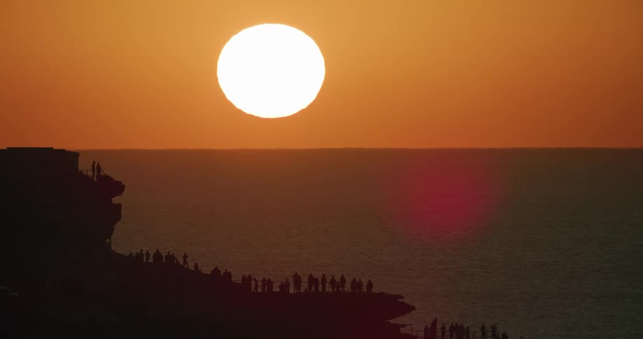 A crowd of people stand along a cliff to watch the sun set