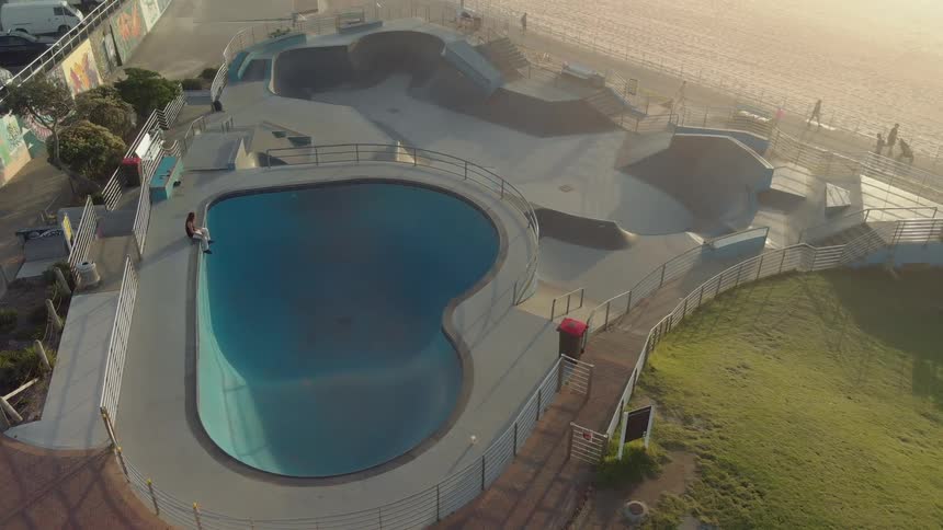 A person sitting on the edge of a pool in an otherwise empty skate park near the beach