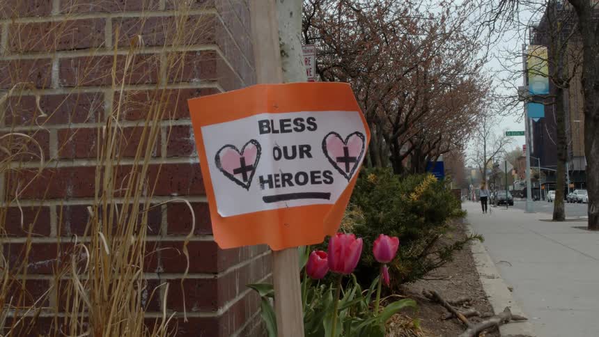 A poster decorated with hearts and crosses says bless our heroes is posted near a brick wall on a city sidewalk