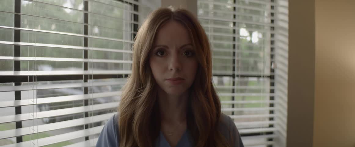 A woman standing next to a window raising her head to stare emotionlessly into the camera