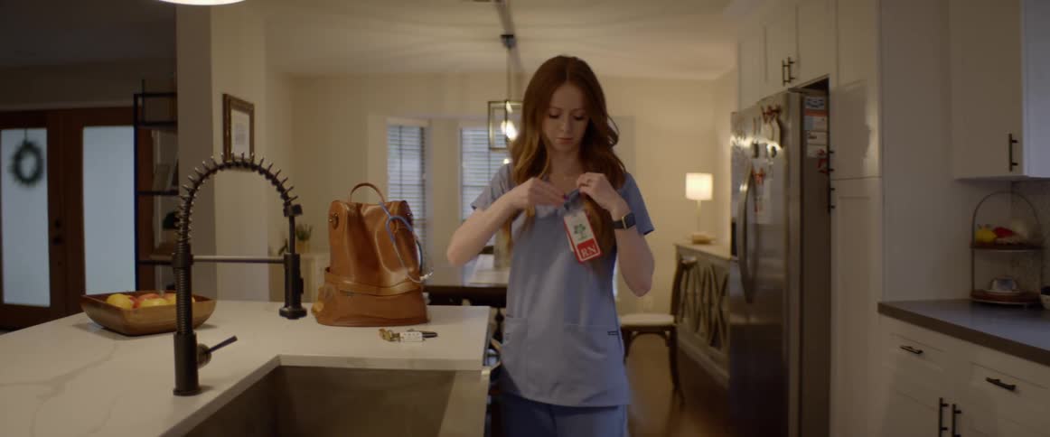 A nurse in blue scrubs gets up from her dining room table and clips on her id picks up her bag and walks out of the kitchen