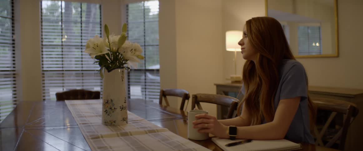 A woman sits at a dining table drinking from a mug