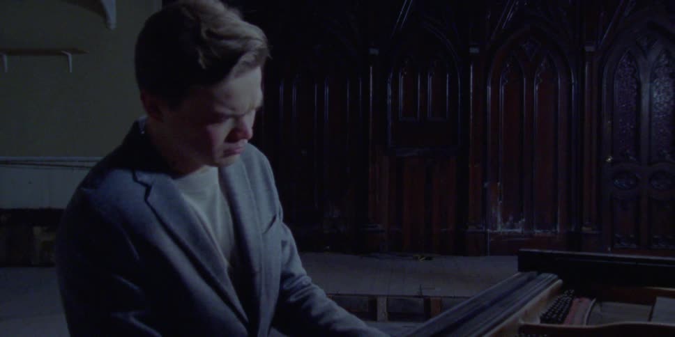A man plays a piano in a dilapidated building with sun shining in through a window