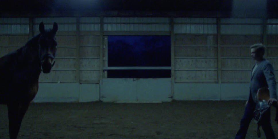 Man steps forward to face horse while carrying a saddle in building near doors
