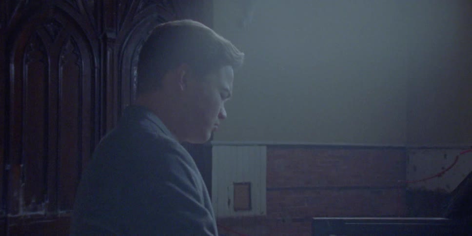 A man sways and plays the piano with his eyes closed in an old run down building