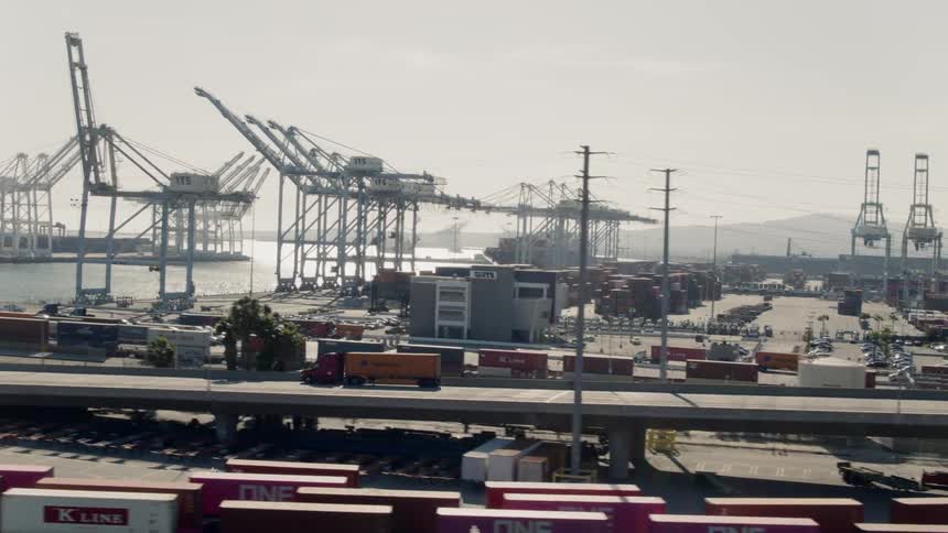 Semi trucks drive down a road that runs alongside a shipping yard by the water