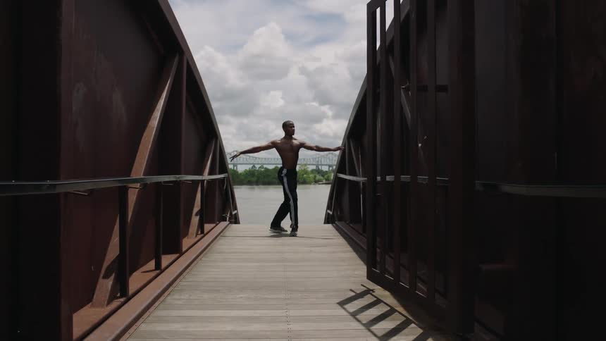 A man on a pier near a lake dancing with a bridge in the background