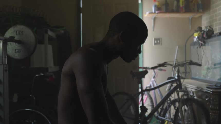 Shirtless black man looking down at the ground raises his head in a dark room