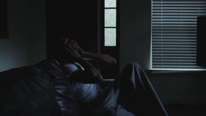 A man sits on his couch and puts his head back in to his hands