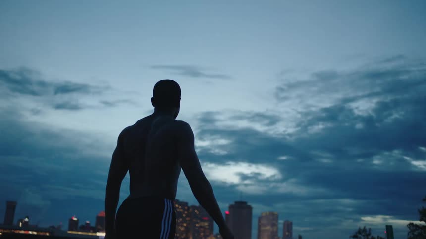 A shirtless man wearing track pants looks over at the city lights as he walks