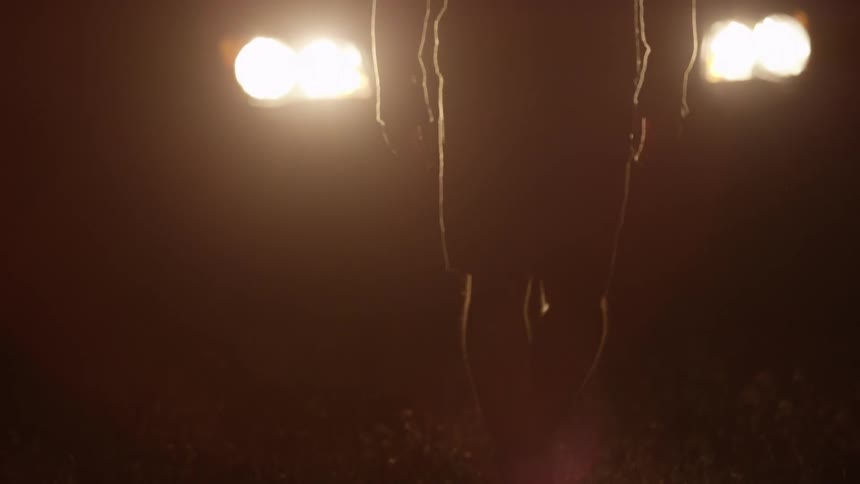 A man walks through grass towards a car's headlights at night