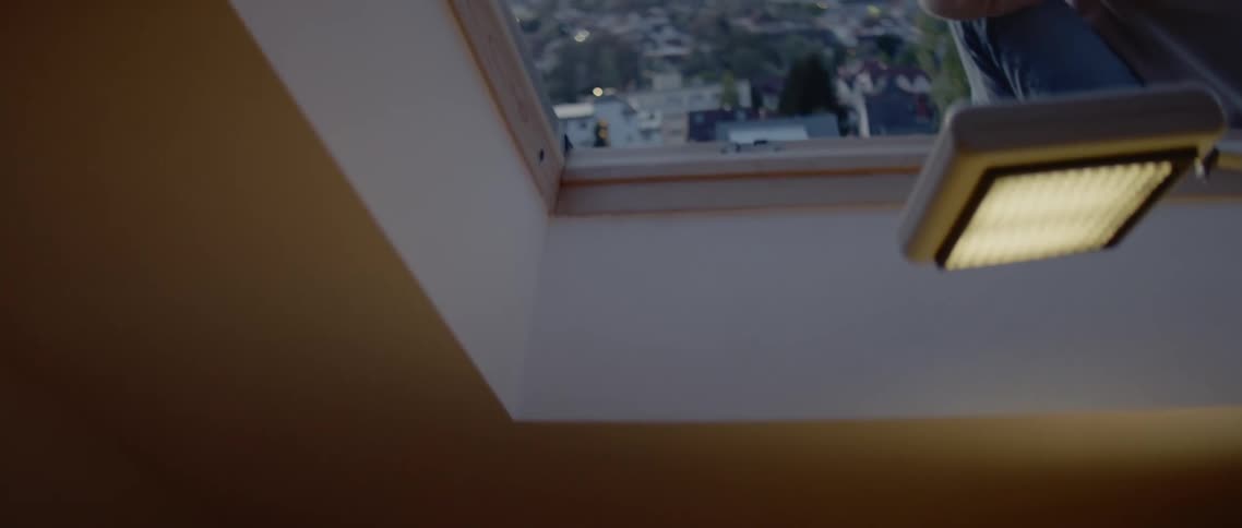 A step ladder sits in a room next to a desk and under an open skylight where a man puts the hood up on his sweatshirt as he looks out over the rooftops at sunrise