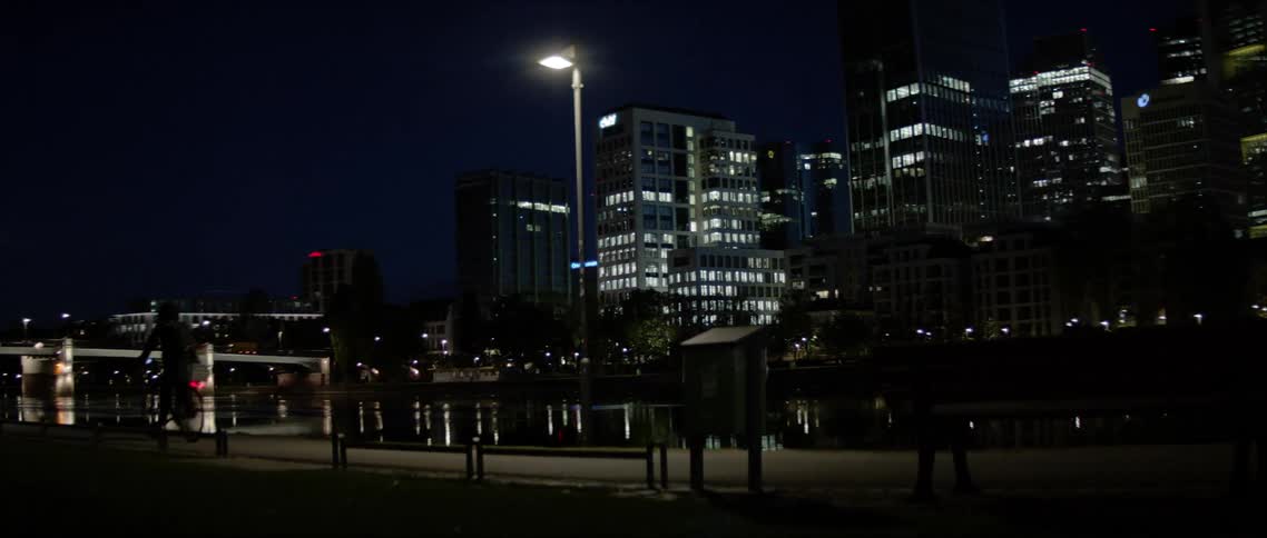 A person rides a bicycle at night on a river side city sidewalk