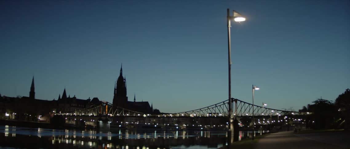 A pathway next to a pond with streetlights with a bridge in the background