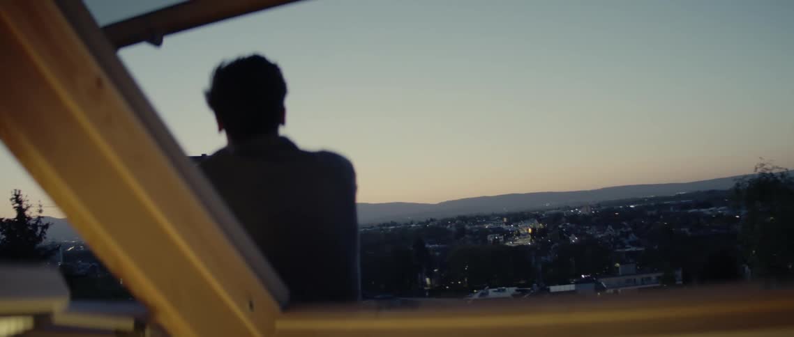 A man sits outside a large open window looking down at the neighborhood lights below at sunset