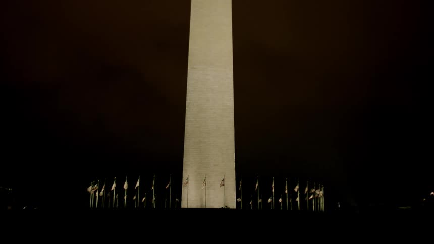 A ring of flags ripple in breeze surrounding a monument at night