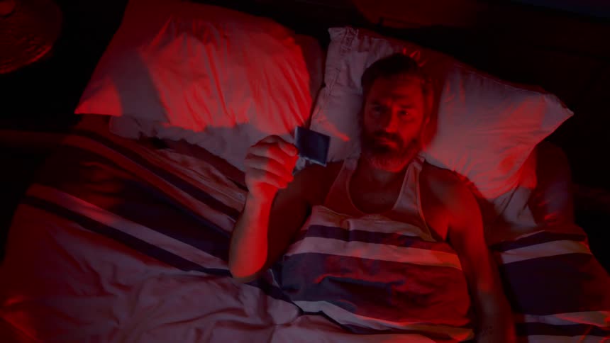 Bearded man lying on the bed looks at a photograph in the dim bedroom