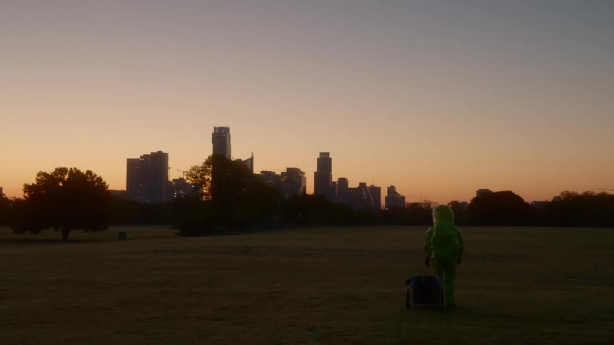 A man in a hazmat suit stands in an open field and looks at the city skyline