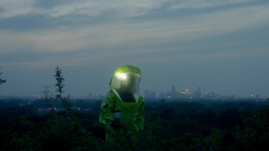 A man wearing a head mounted flashlight in a yellow hazmat suit standing on a hill outside a metropolis