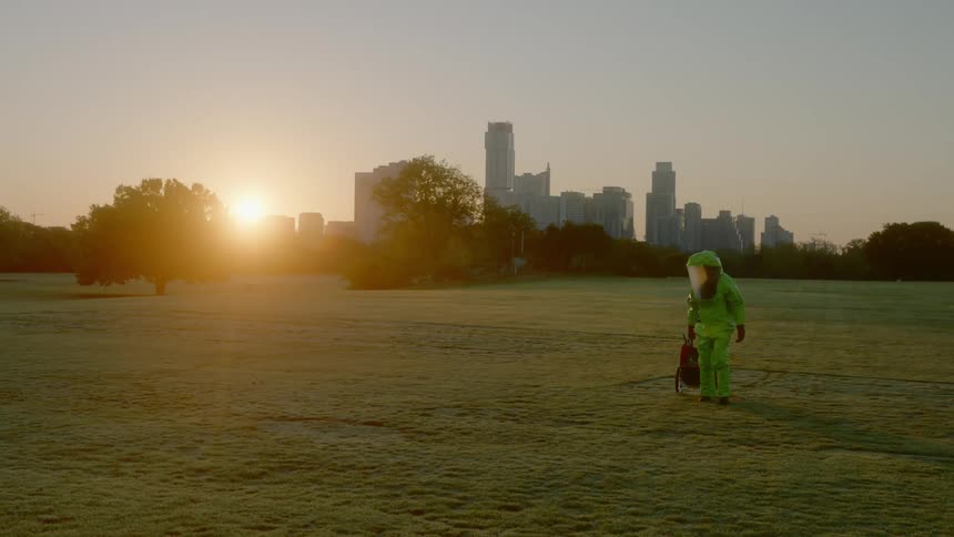 A man in a full green hazmat suit walks in an empty public park wheeling a cart behind him with the city skyline above the trees